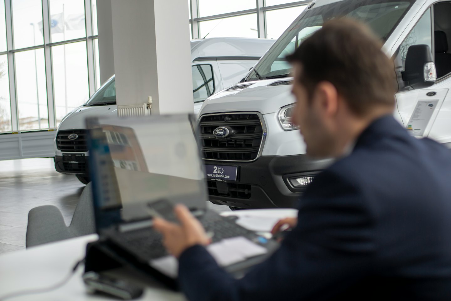 Person types on laptop at desk in car showroom with vans.